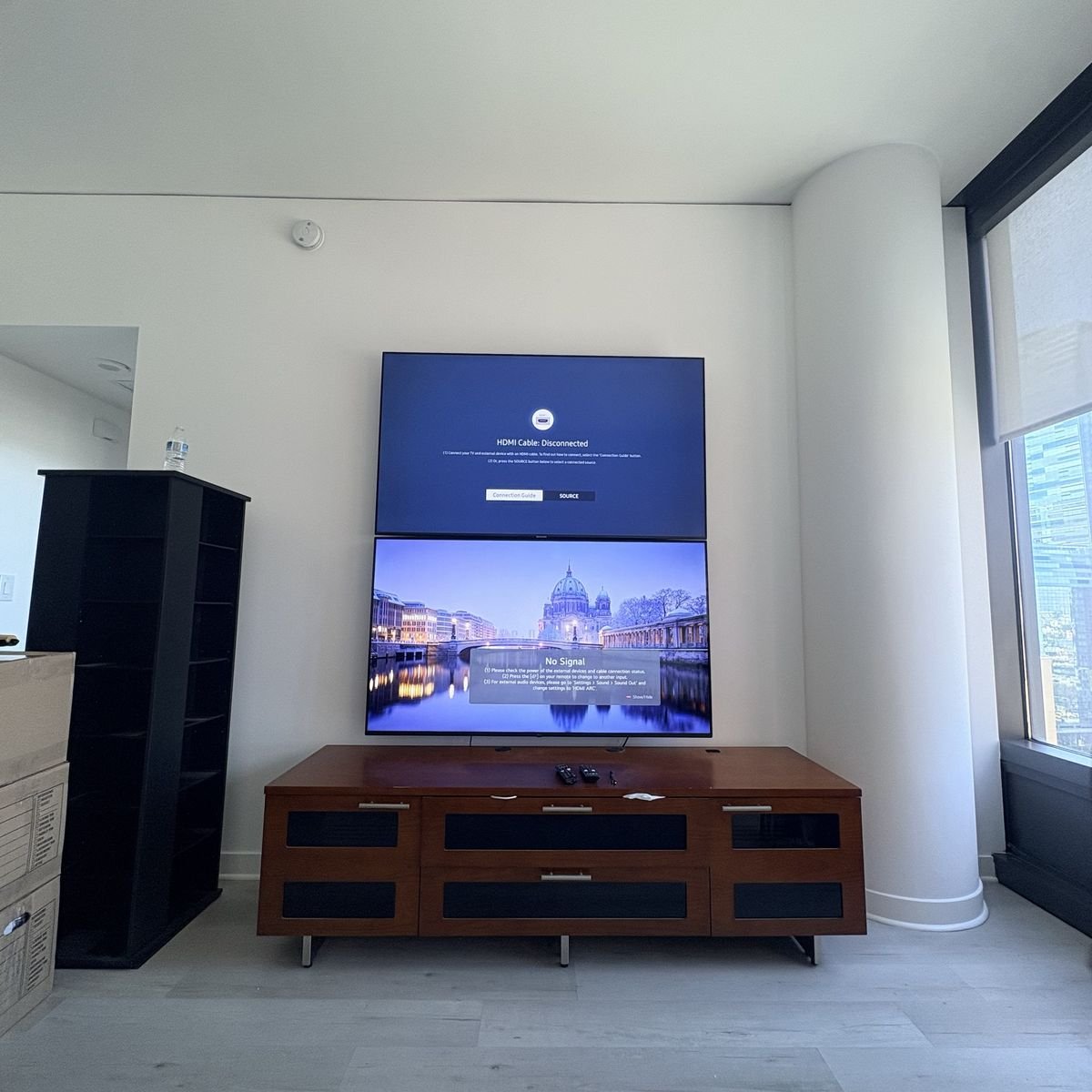 Two TVs stacked on the wall above a media console in a high-rise apartment